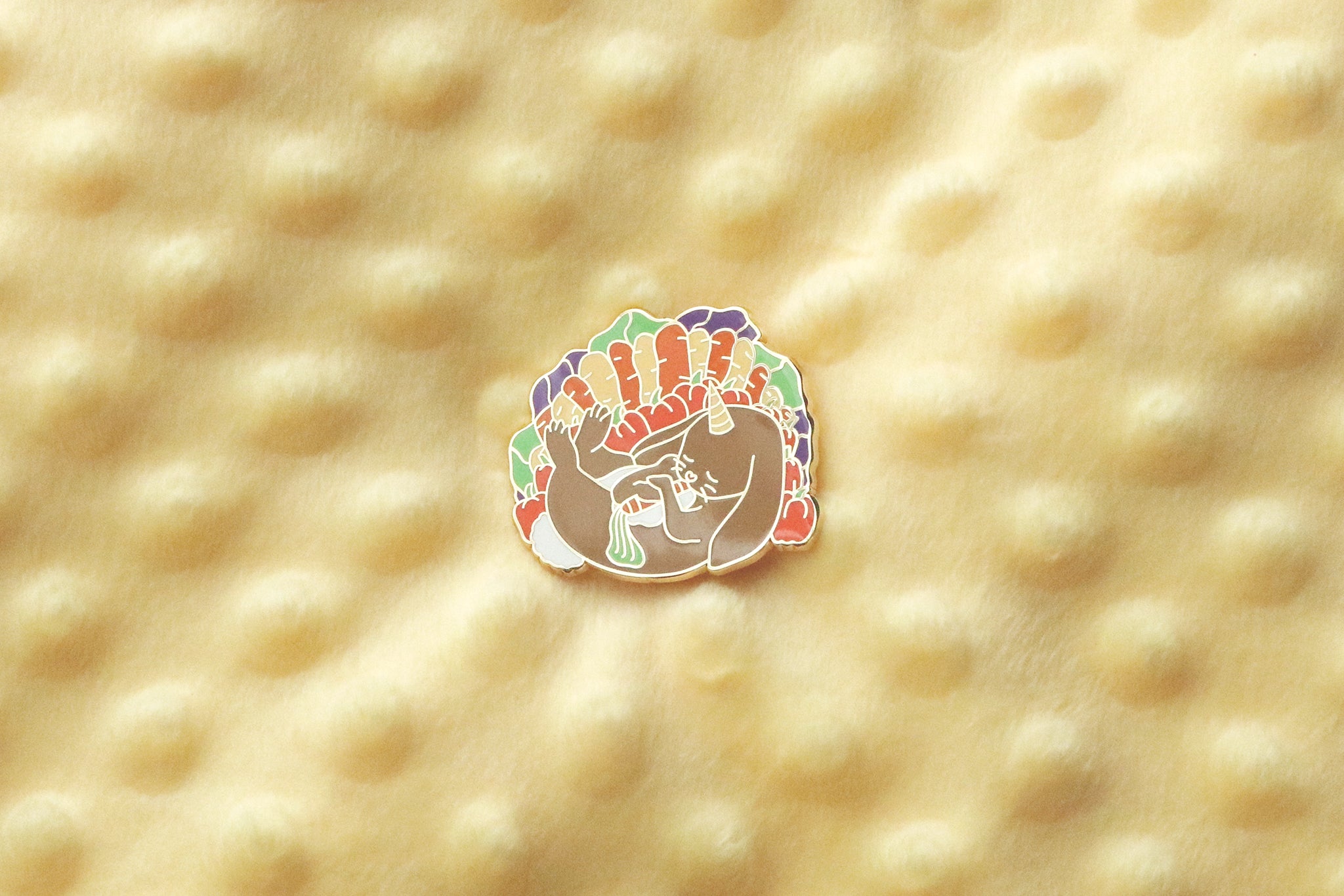 A gold plated enamel pin depicting a cuddly almiraj surrounded by her favorite vegetable snacks of cabbage, carrots, and bell peppers.