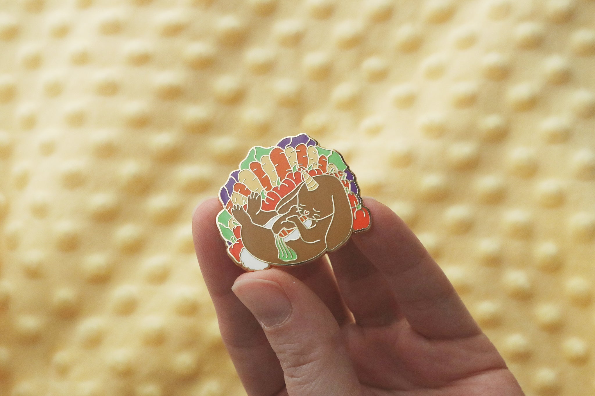 A hand holding a gold plated enamel pin depicting a cuddly almiraj surrounded by her favorite vegetable snacks of cabbage, carrots, and bell peppers.