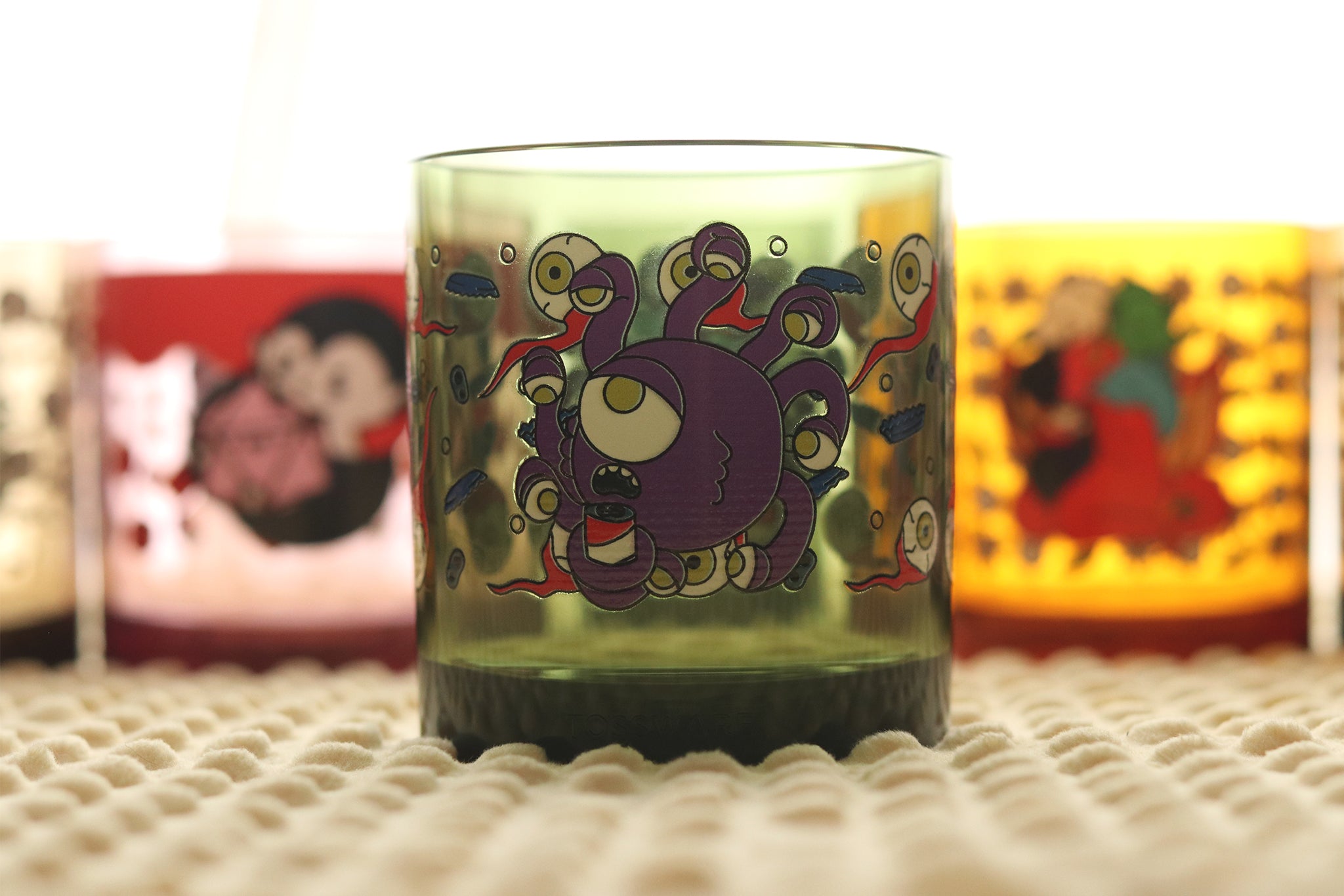 Beholder Beer Holder Tossware glass featuring purple D&D beholder monster with multiple eyestalks holding a beer can on a green tumbler photographed on textured surface