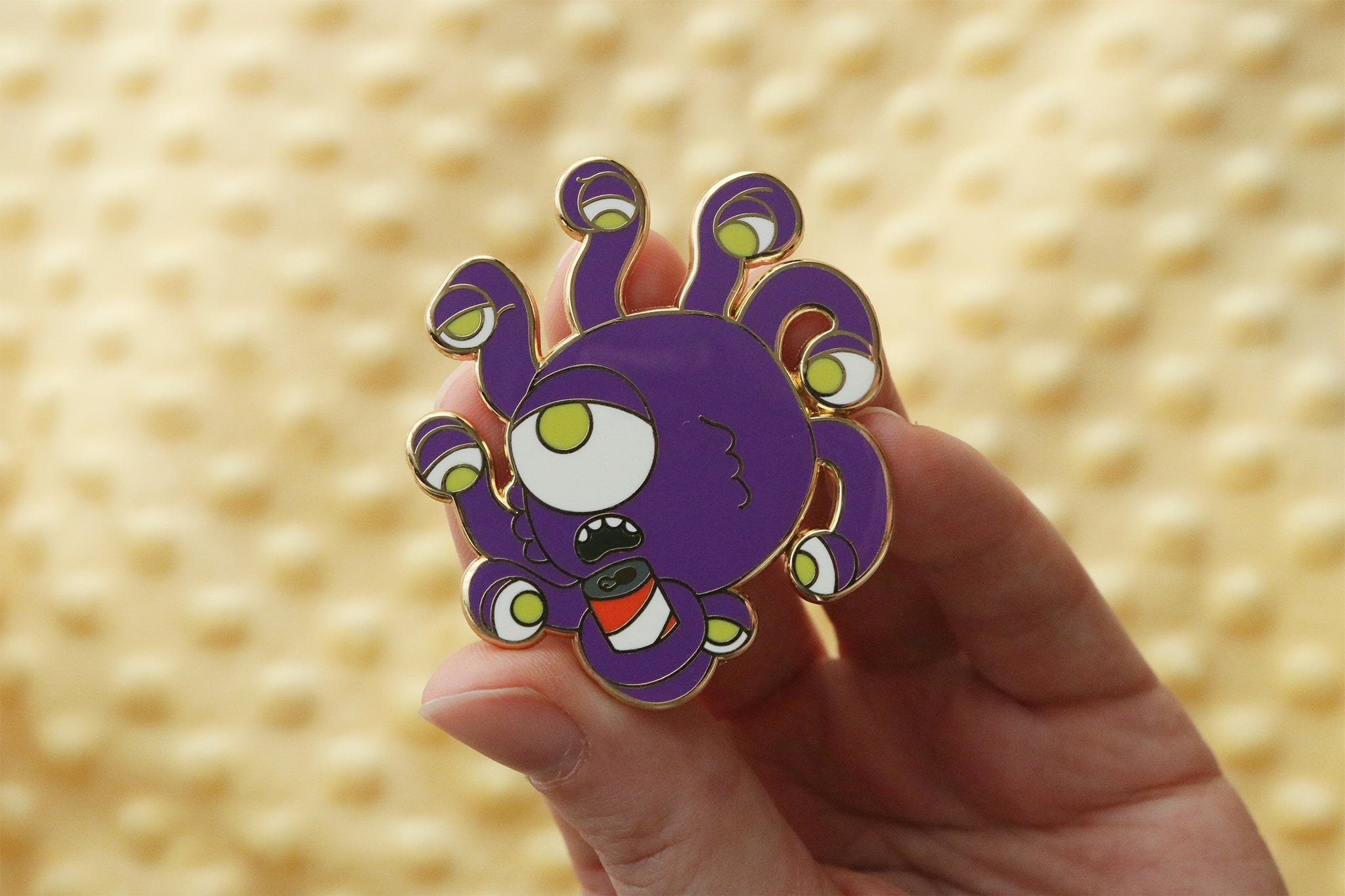 Enamel pin featuring a cute cartoon beholder monster designed with a purple spherical body, multiple eye stalks with green eyes, and holding a small red beer can.