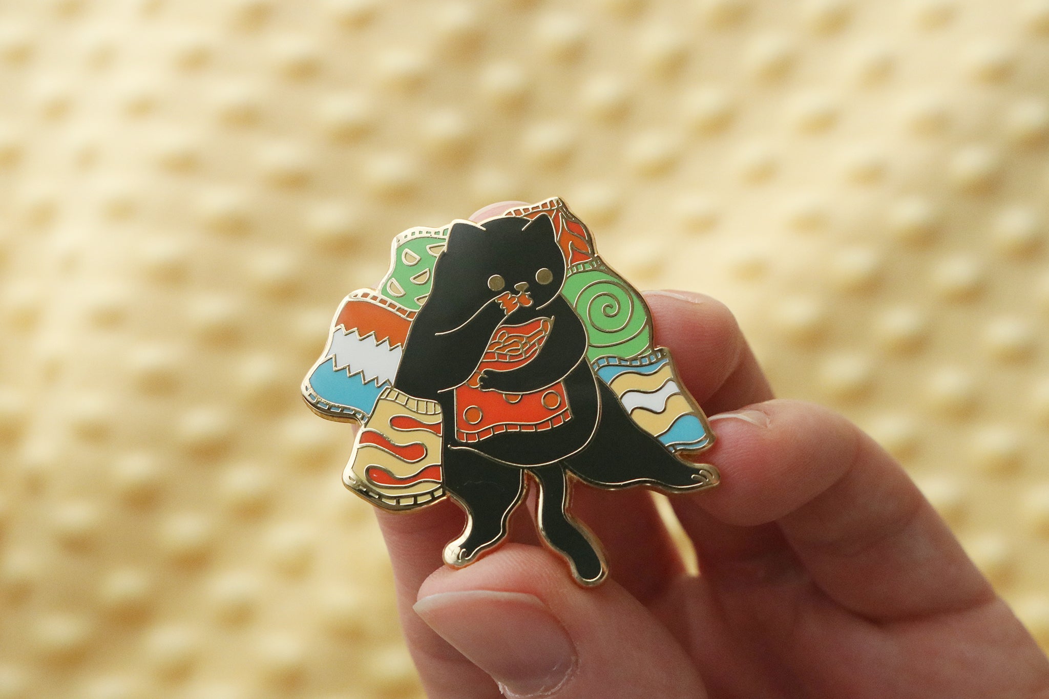 Hand holding an enamel pin featuring a black cat silhouette surrounded by bags of colorful potato chips in vibrant colors of green, orange, blue, and red
