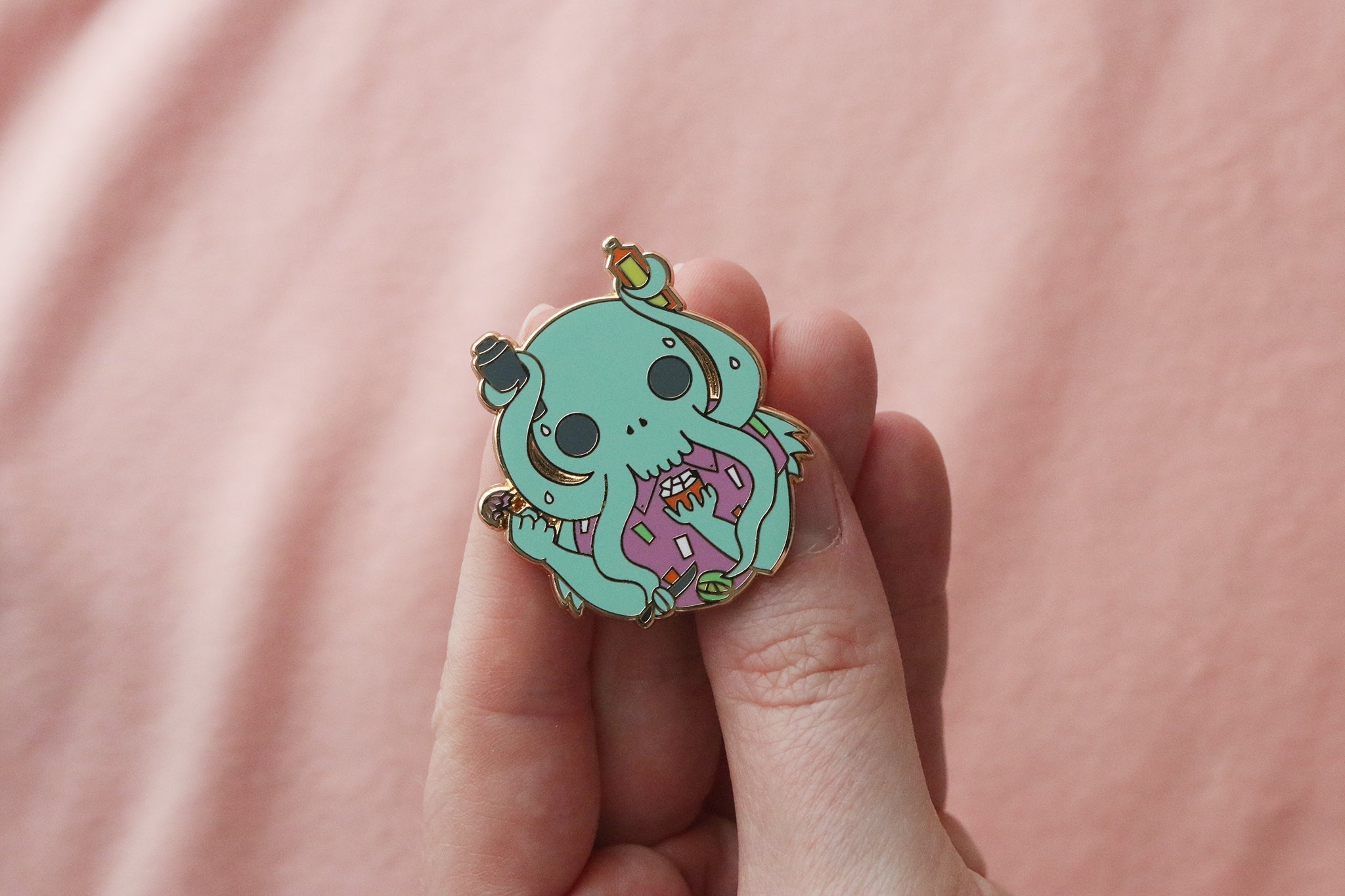 An enamel pin held in a hand showing a cute, cartoonish Cthulhu character in mint green with large gray eyes and tentacles holding cocktail tools.