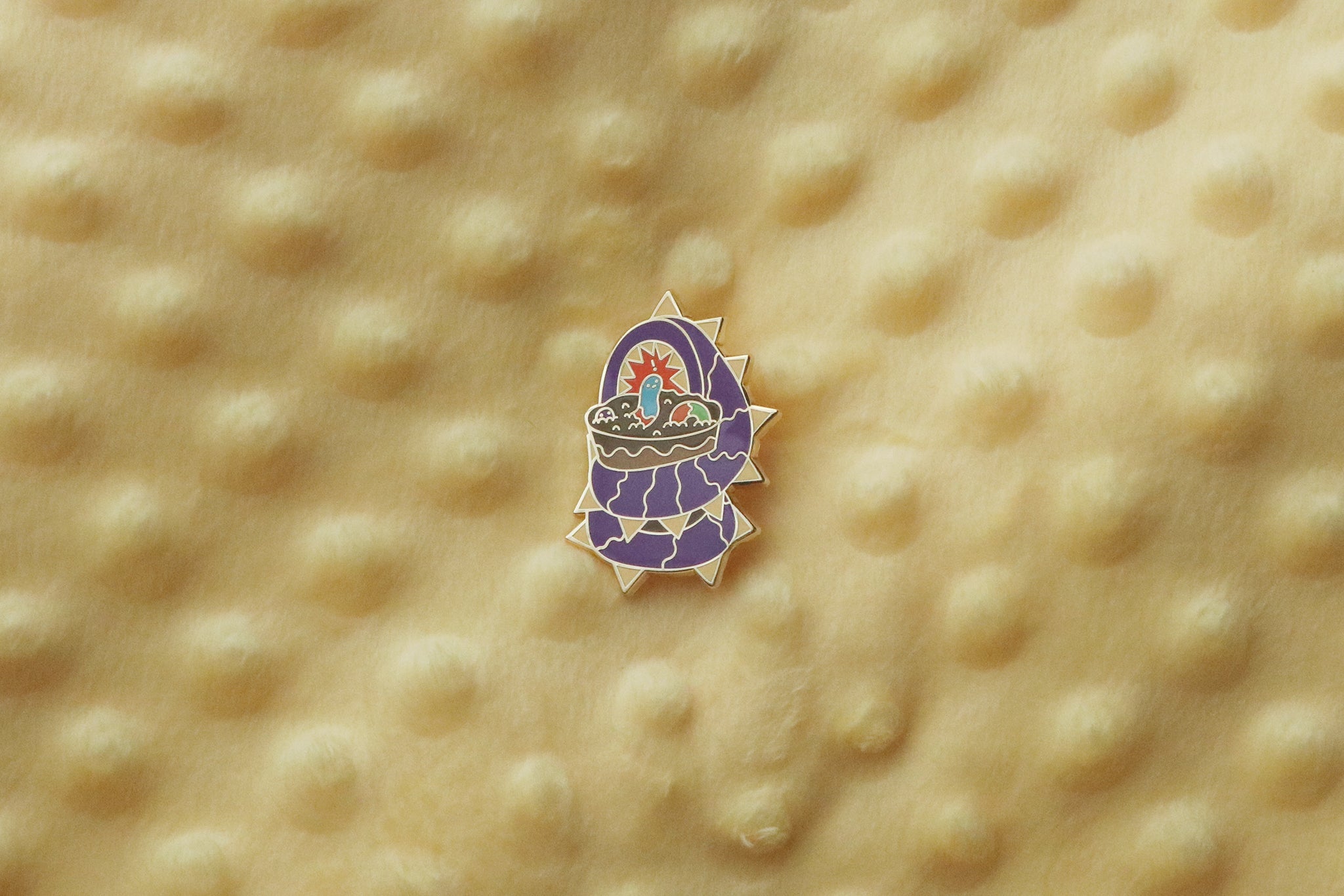 Enamel pin photographed on a textured yellow fabric background showing a large purple segmented worm creature with spiky protrusions coiled around a dirt cup dessert. The cup contains chocolate cookie crumbs and pudding with colorful gummy worms.