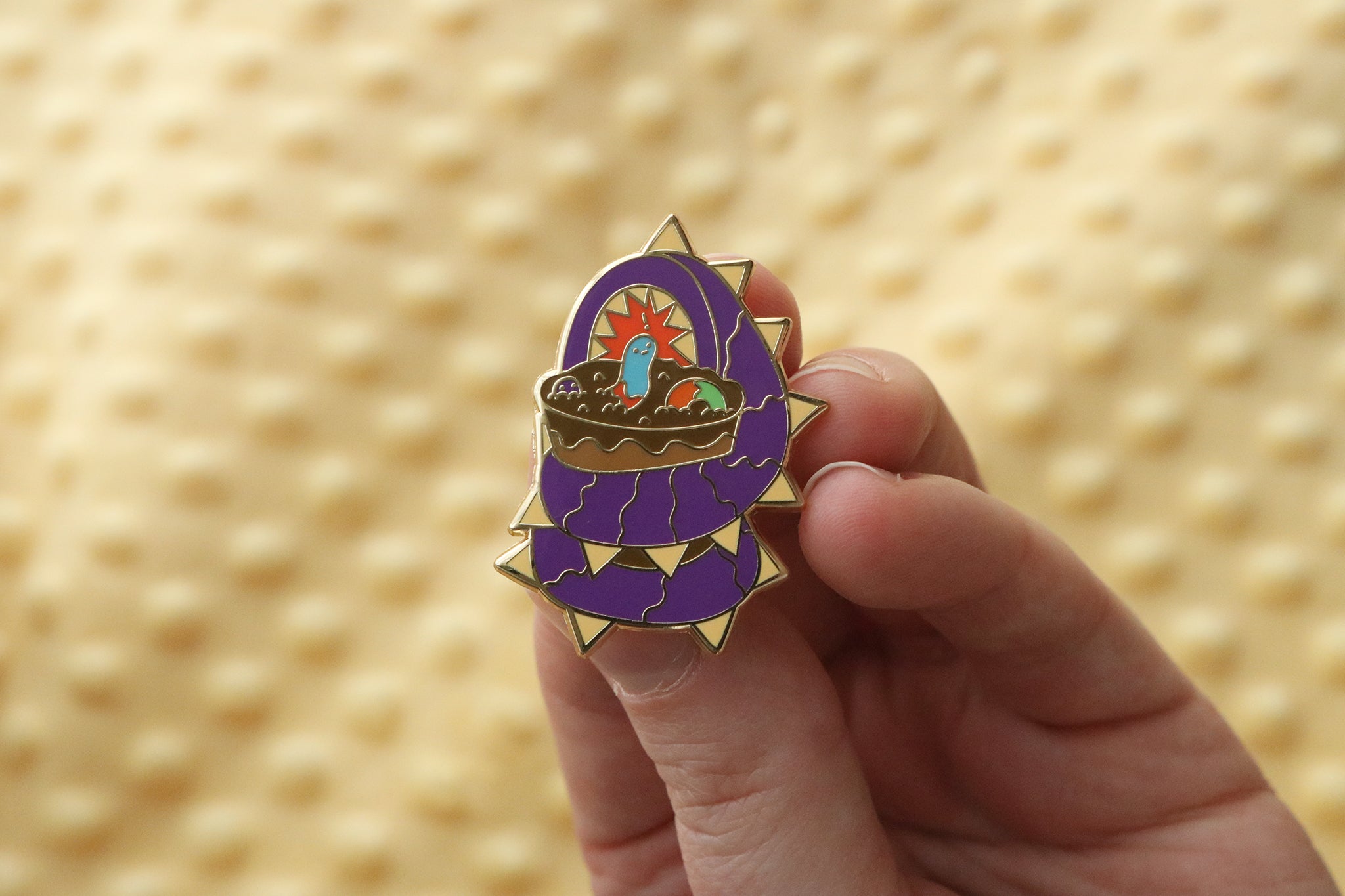 Close-up of an enamel pin held between fingers showing a purple worm monster with golden spikes wrapped around a dirt cup dessert. The cup features chocolate crumb 'dirt' with small multicolored gummy worms. The pin has detailed enamel work with gold-toned metal accents, photographed against a yellow textured background.