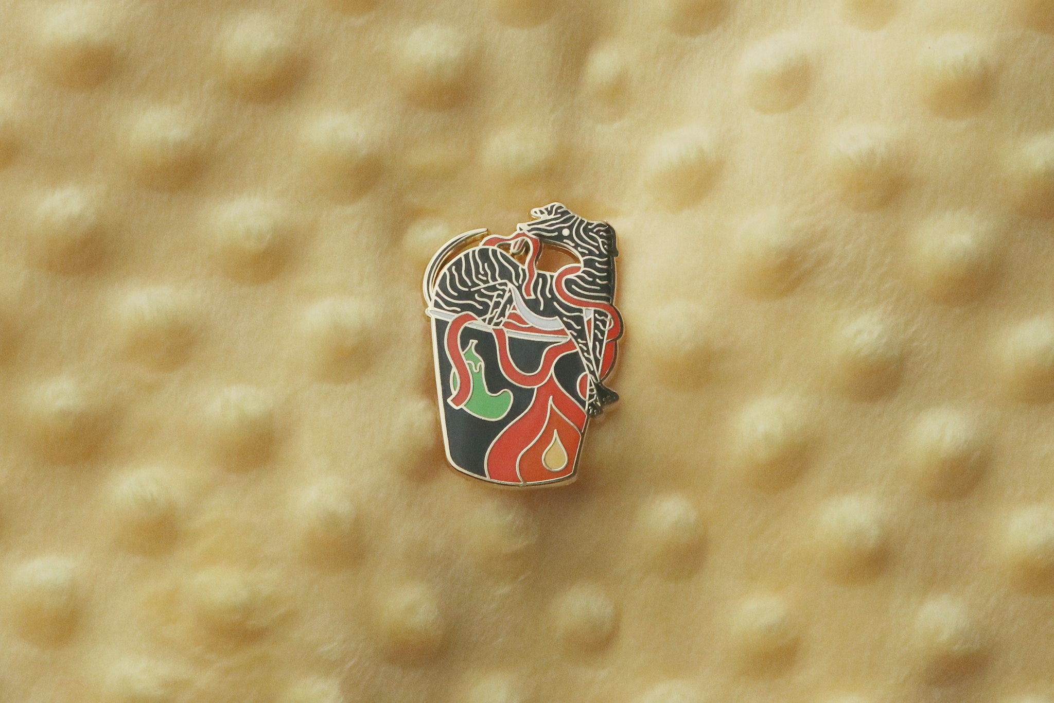 A black hell hound enamel pin photographed flat against a yellow background. The pin depicts a demon-like canine creature laying in a black cup of flaming red noodles. The design combines instant ramen imagery with mythological hellhound aesthetics, finished with gold metal plating.
