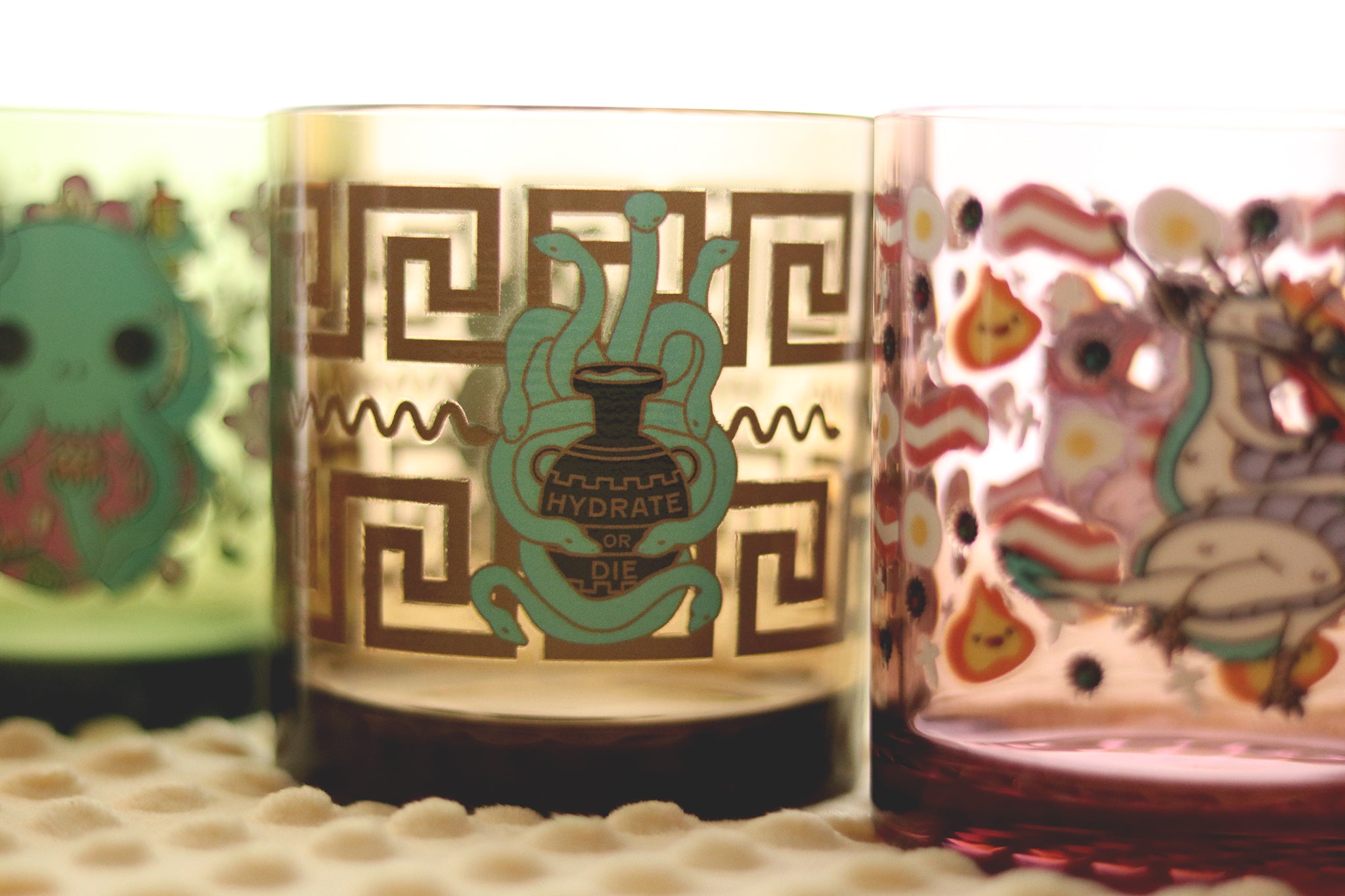 Close-up view of Hydrate or Die drinking glass showing detailed artwork of multi-headed hydra serpent embracing ancient water vessel, displayed alongside other colorful Tossware cups.