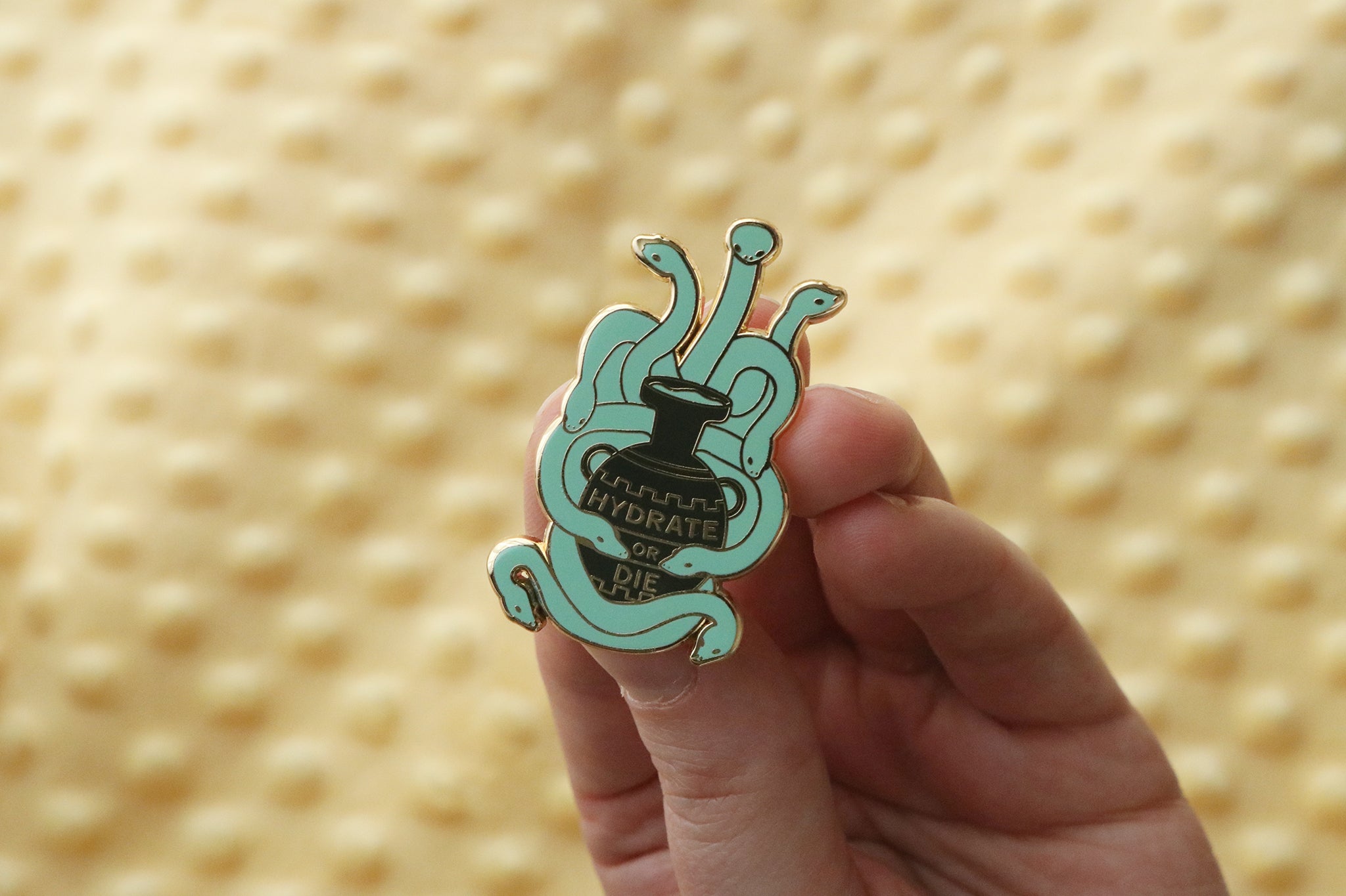 A teal hydra enamel pin held in hand, showing the mythological multi-headed serpent creature coiled around a black water vessel inscribed with 'HYDRATE OR DIE.' The design cleverly plays on the hydra's association with water and modern hydration culture.