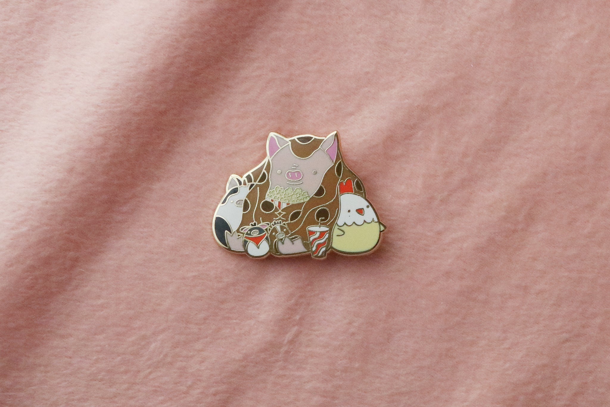 Enamel pin featuring a pig wrapped in a brown blanket, holding a bowl of popcorn with a striped drink cup nearby, surrounded by cute farm animal plushies including a cow, sheep, and chicken.