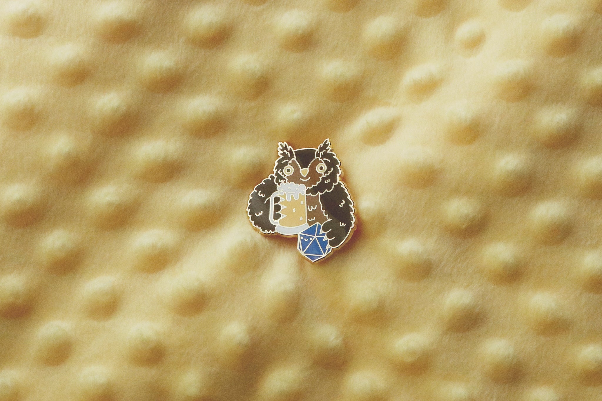 Enamel pin on a yllow textured fabric background showing a cute brown owlbear character. The creature is holding a foamy beer mug in one hand and a blue twenty-sided die (d20) in the other. The pin features detailed enamel work with gold-toned metal accents.