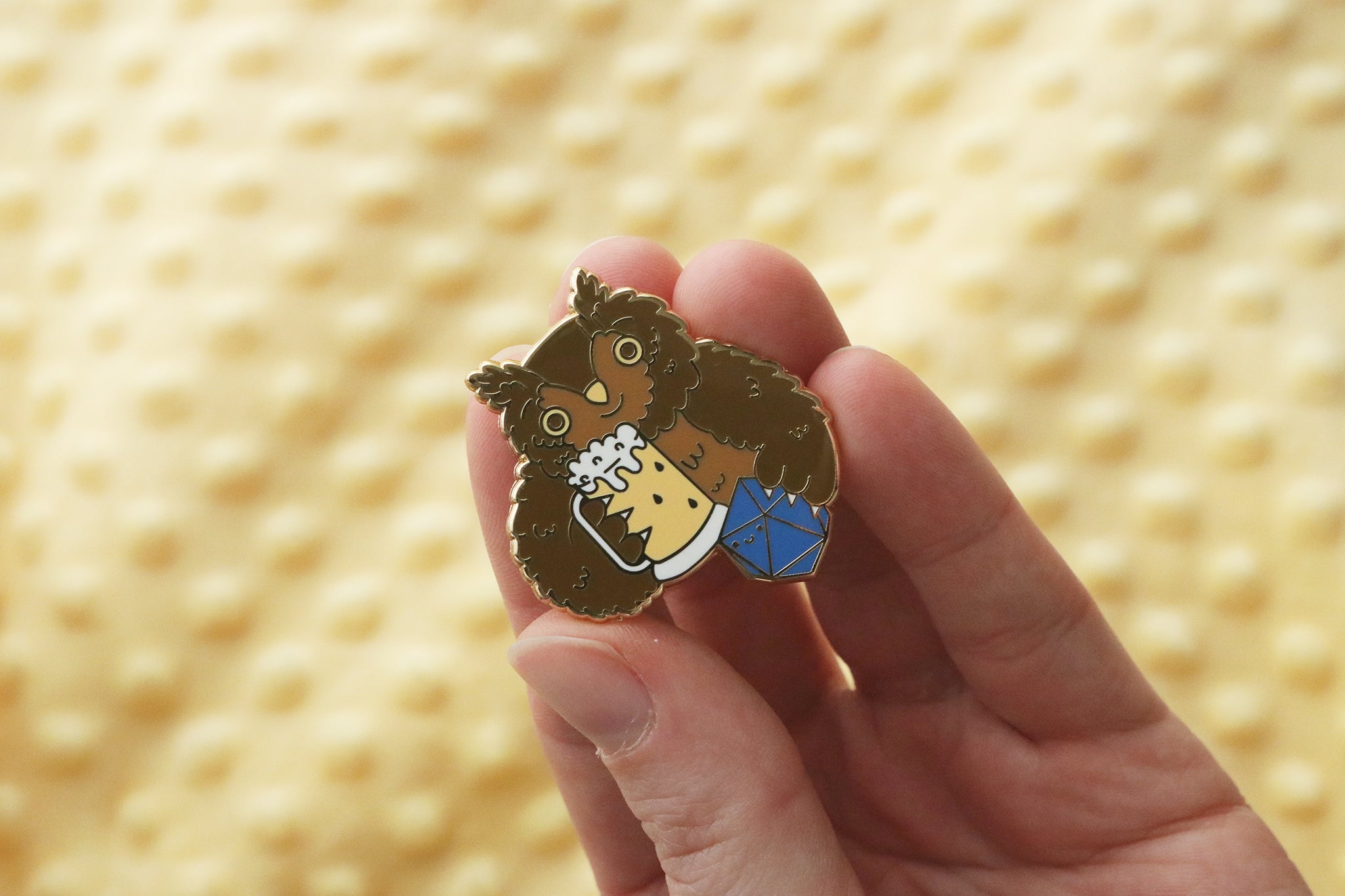 Close-up of an enamel pin held between fingers depicting a friendly owlbear with large round eyes and fuzzy brown fur. The character grips a beer mug with white foam on top in one paw and a blue d20 gaming die in the other. The pin shows intricate enamel detailing against a warm yellow background, combining D&D gaming elements with a cheerful drinking theme