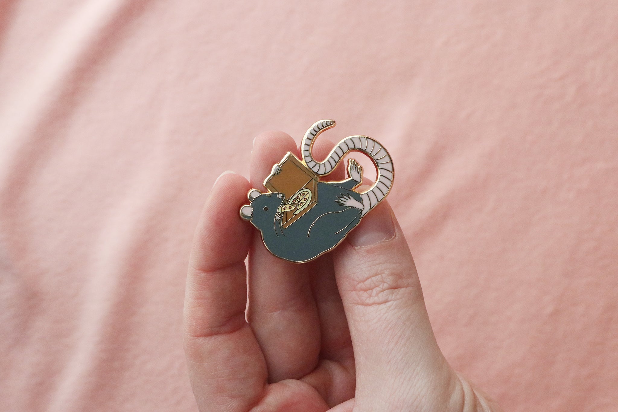 Enamel pin held between fingers showing a large gray rat creature with small ears and a long striped tail, holding and eating a slice of pepperoni pizza out of the delivery box. The pin features gold-toned accents and is photographed against a soft pink fabric background