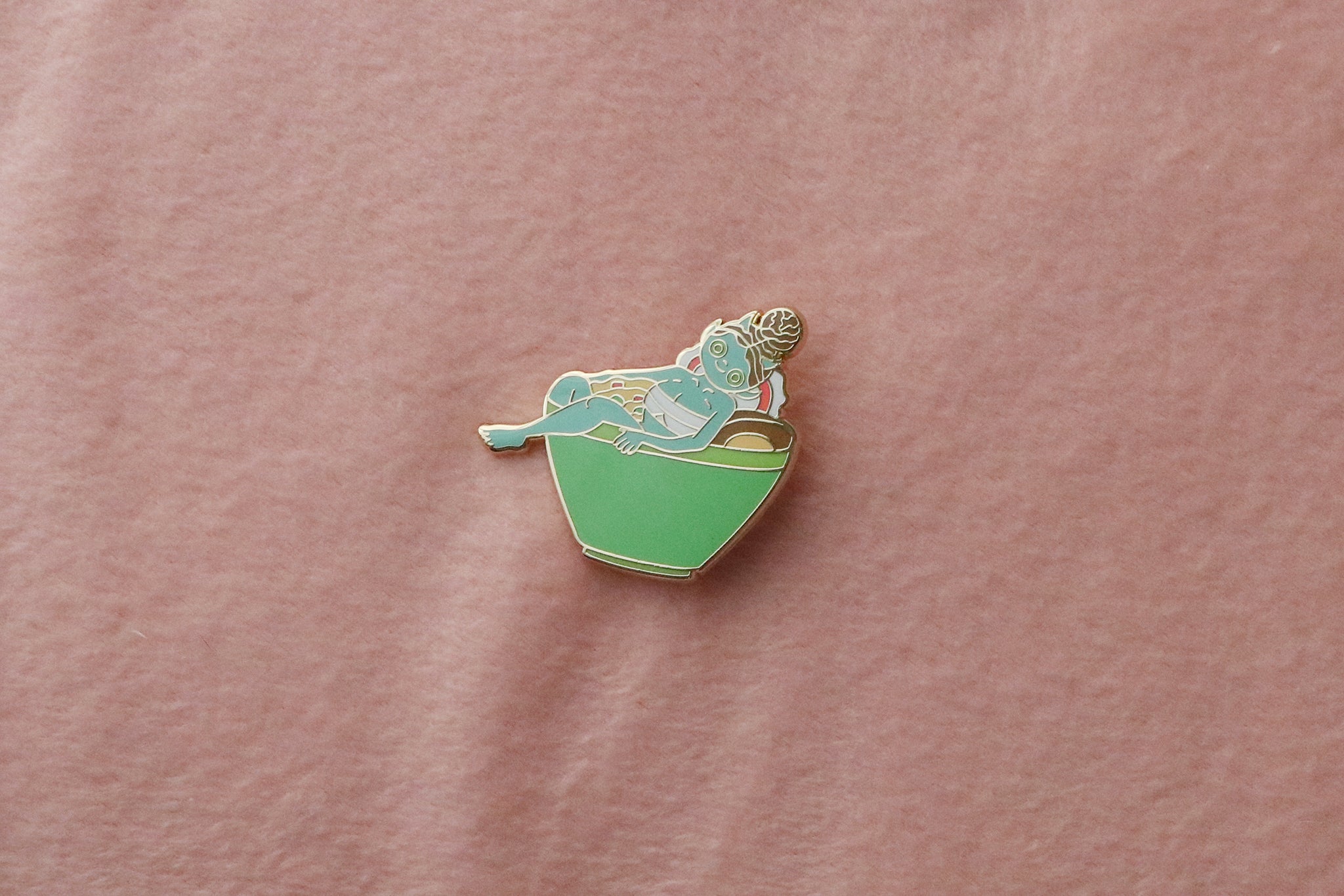 Enamel pin on pink fabric showing a blue-skinned oni monster girl with small horns and brown curly hair lounging relaxedly in a bright green ramen bowl. She's partially submerged in the noodle broth with ramen noodles visible around her. The pin features gold-toned metal accents and detailed enamel work.