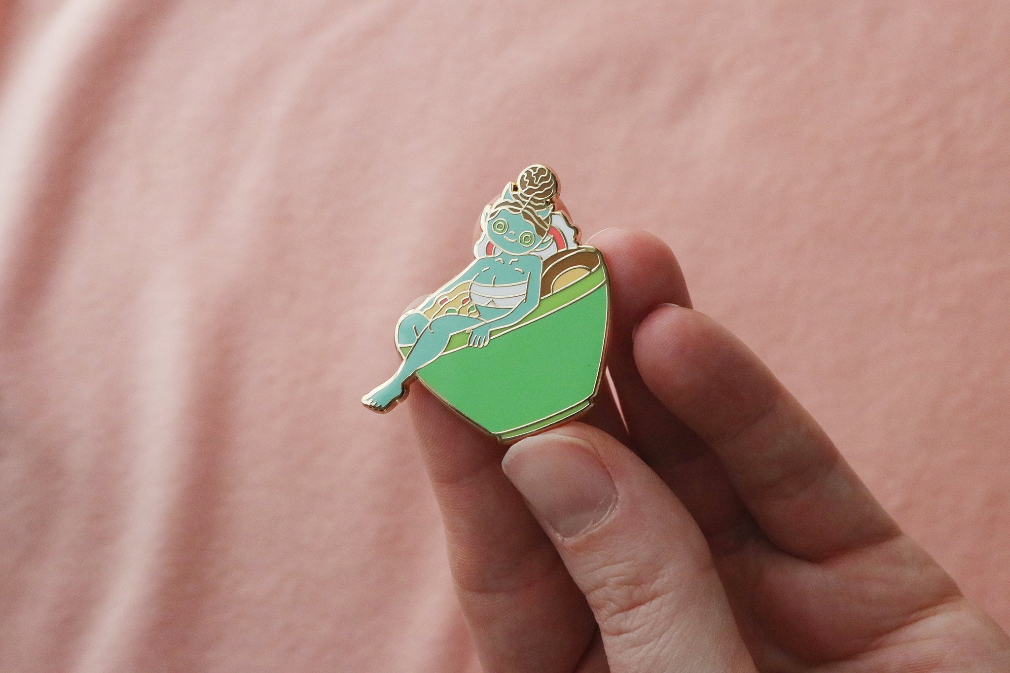 Close-up of an enamel pin held between fingers depicting a cute blue oni girl with horns and light hair reclining in a green ramen bowl filled with noodles and broth. The character has a relaxed, content expression while soaking in the bowl. The pin shows intricate enamel detailing with metallic gold plating, photographed against a soft pink background