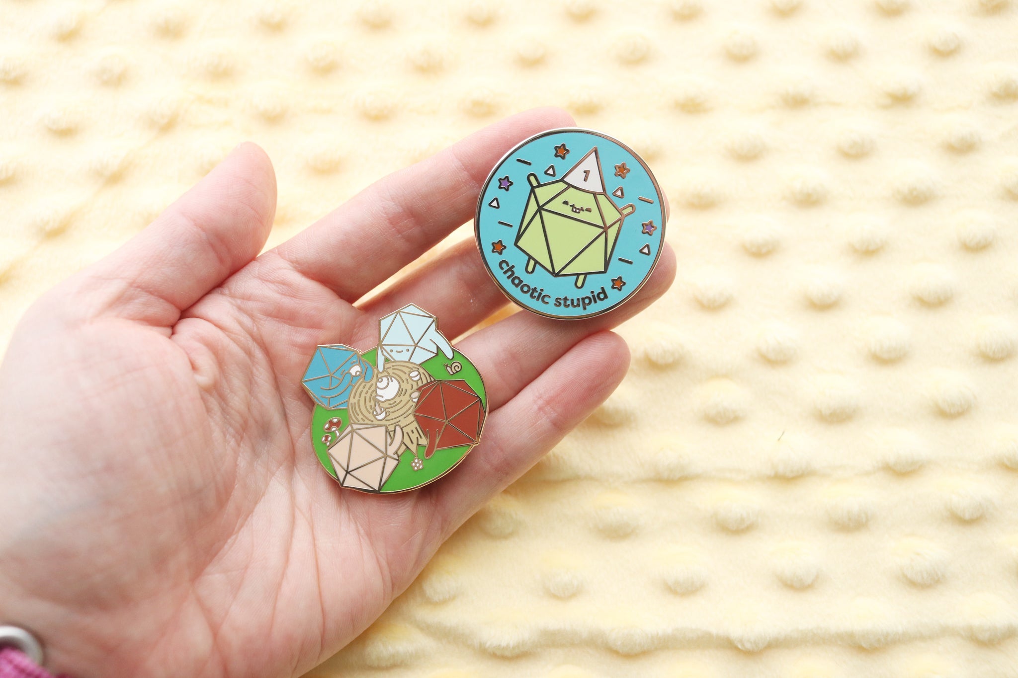 Hand holding two enamel pins. One pin is a circular teal pin that reads Chaotic Stupid. There is a lime green d20 wearing a dunce cap with the number 1 on it. Stars, triangles, and dashes surround the dice.