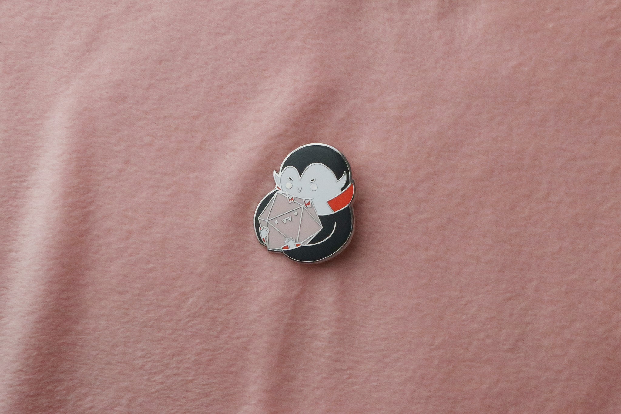 Hard enamel pin of kawaii-style vampire character with black hair and red collar, holding geometric D20 gaming die