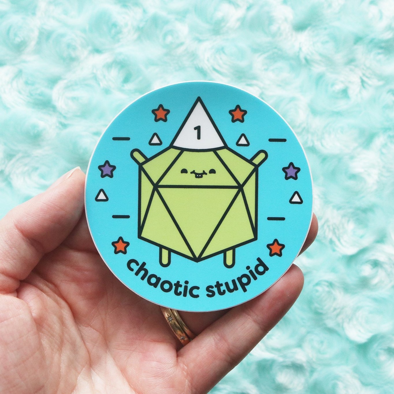 Sticker photographed on blue fur background. Sticker reads "Chaotic Stupid." A lime green d20 wears a white dunce cap with the number 1 on it.