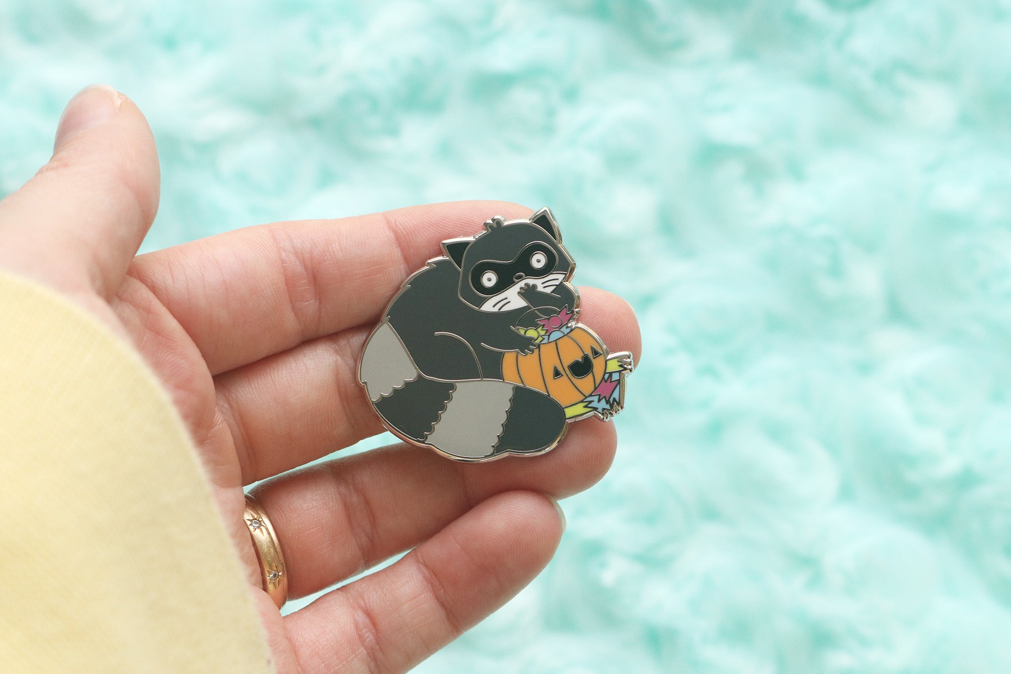 Raccoon enamel pin. Fluffy critter is caught red handed eating trick-or-treat candy out of an orange pumpkin basket.