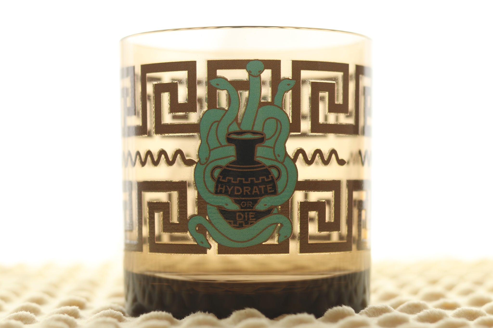 Hydrate or Die Tossware glass featuring teal mythological hydra with multiple heads wrapped around Greek amphora with 'Hydrate or Die' text, surrounded by geometric Greek key pattern border, photographed on textured surface.