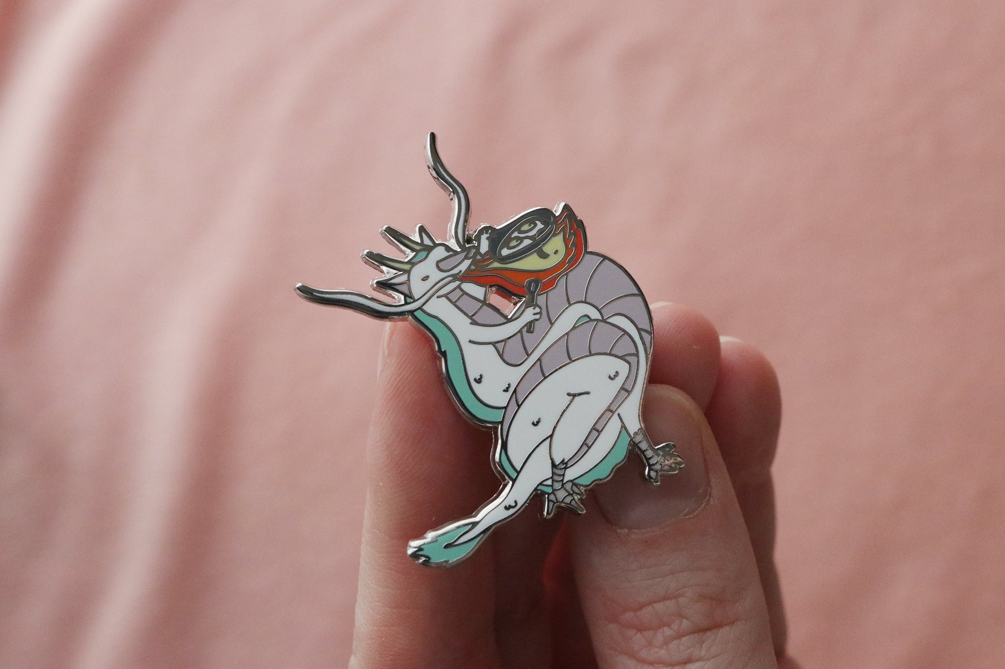 An enamel pin held in hand showing a white and mint green dragon breathing orange and red flames to cook a breakfast of eggs in a gray frying pan. The dragon has a cute, friendly expression with small eyes and is depicted in a kawaii art style. The pin features gold metal plating and is photographed against a soft pink background