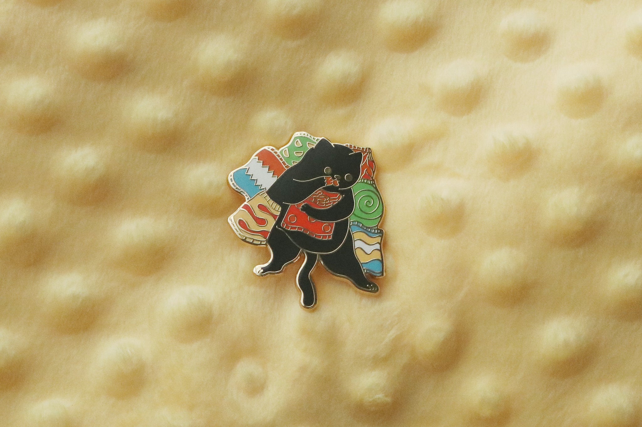 Enamel pin featuring a black cat munching on cheesy puffs while surrounded by colorful snack foods.
