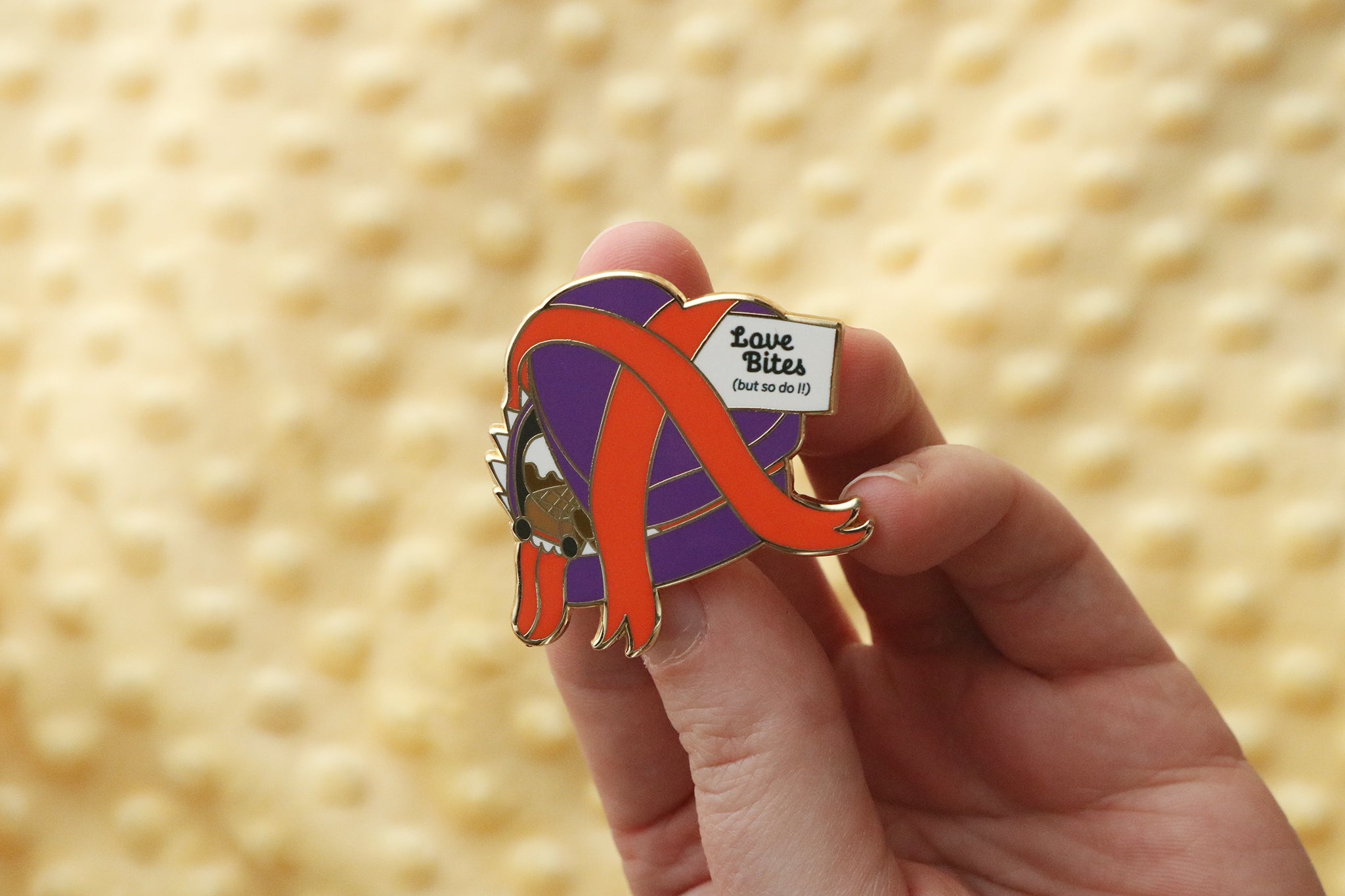 Hand holding a Heart-shaped chocolate mimic box enamel pin in purple and red with decorative ribbons and text reading 'Love Bites (but so do I)' 