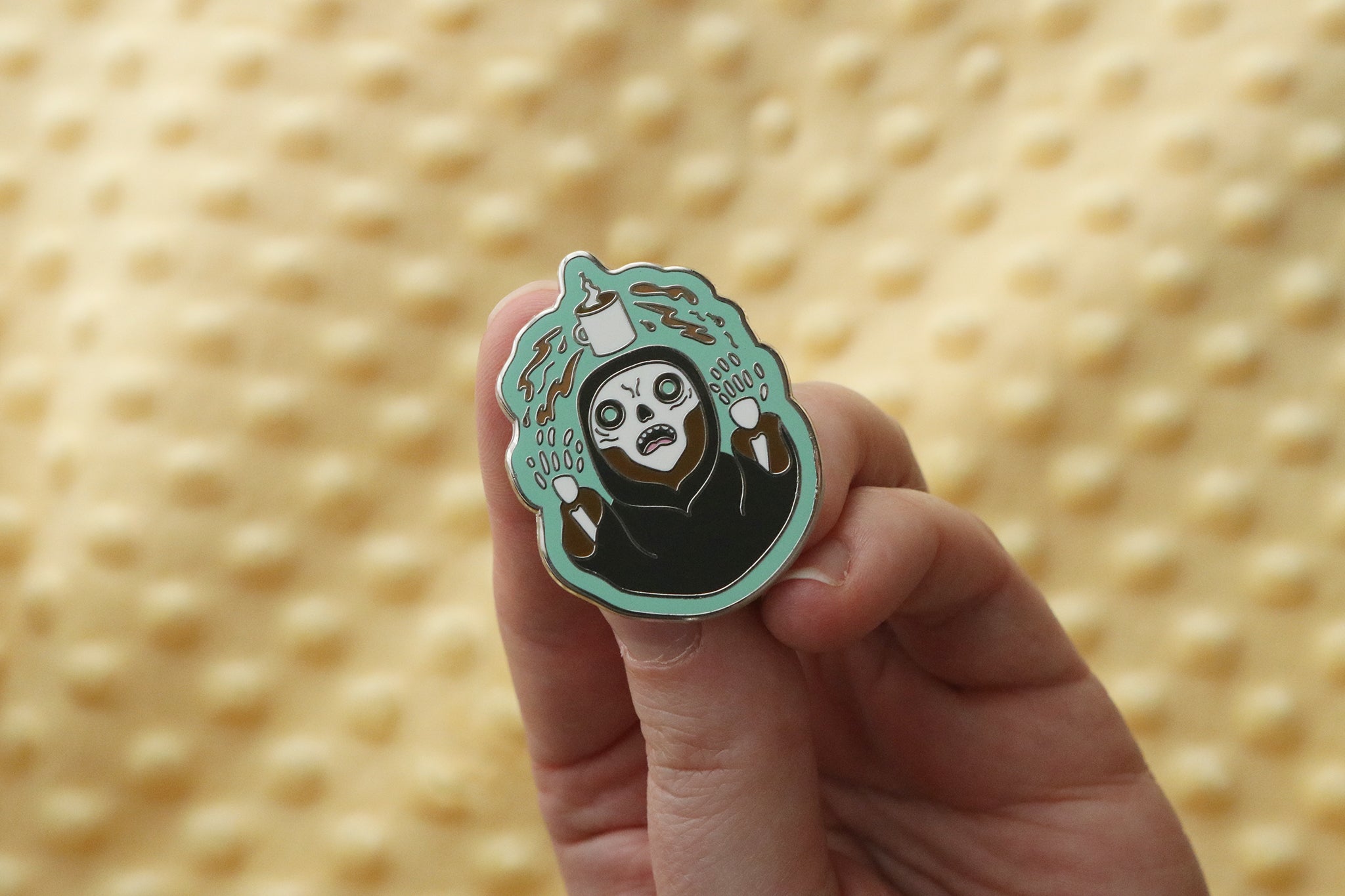 Hard enamel pin with teal background showing a hooded skeletal figure in a black robe with teal glowing eyes, holding up hands in a summoning gesture. Wisps of coffee colored magic flow out of his fingers as a white coffee mug steams above his head.