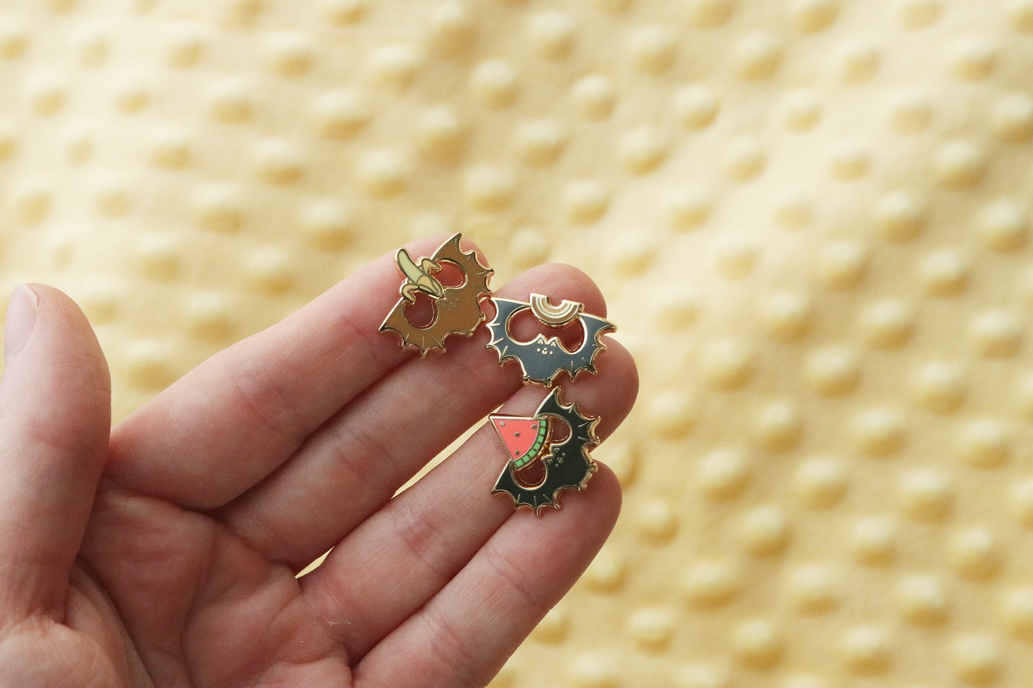Three enamel pins of fruit bats holding their favorite fruit snacks. 