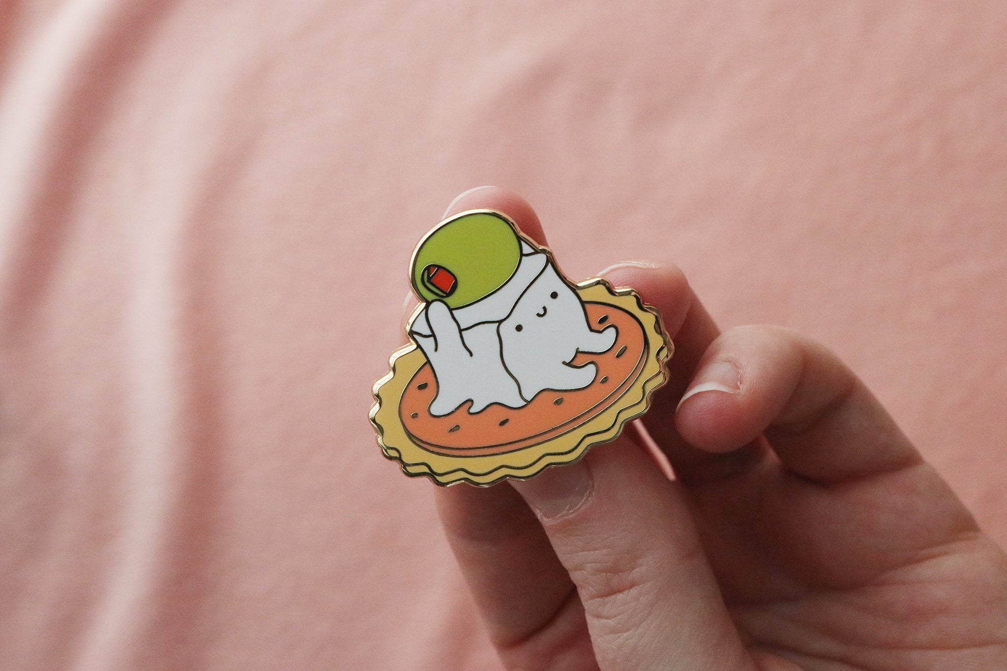 A cheese cube enamel pin held in hand, showing the adorable white gelatinous cube monster with dot eyes and a small smile, perched on a circular cracker decorated with pepperoni. The cube figure is raising a green stuffed olive overhead in a cheerful pose.