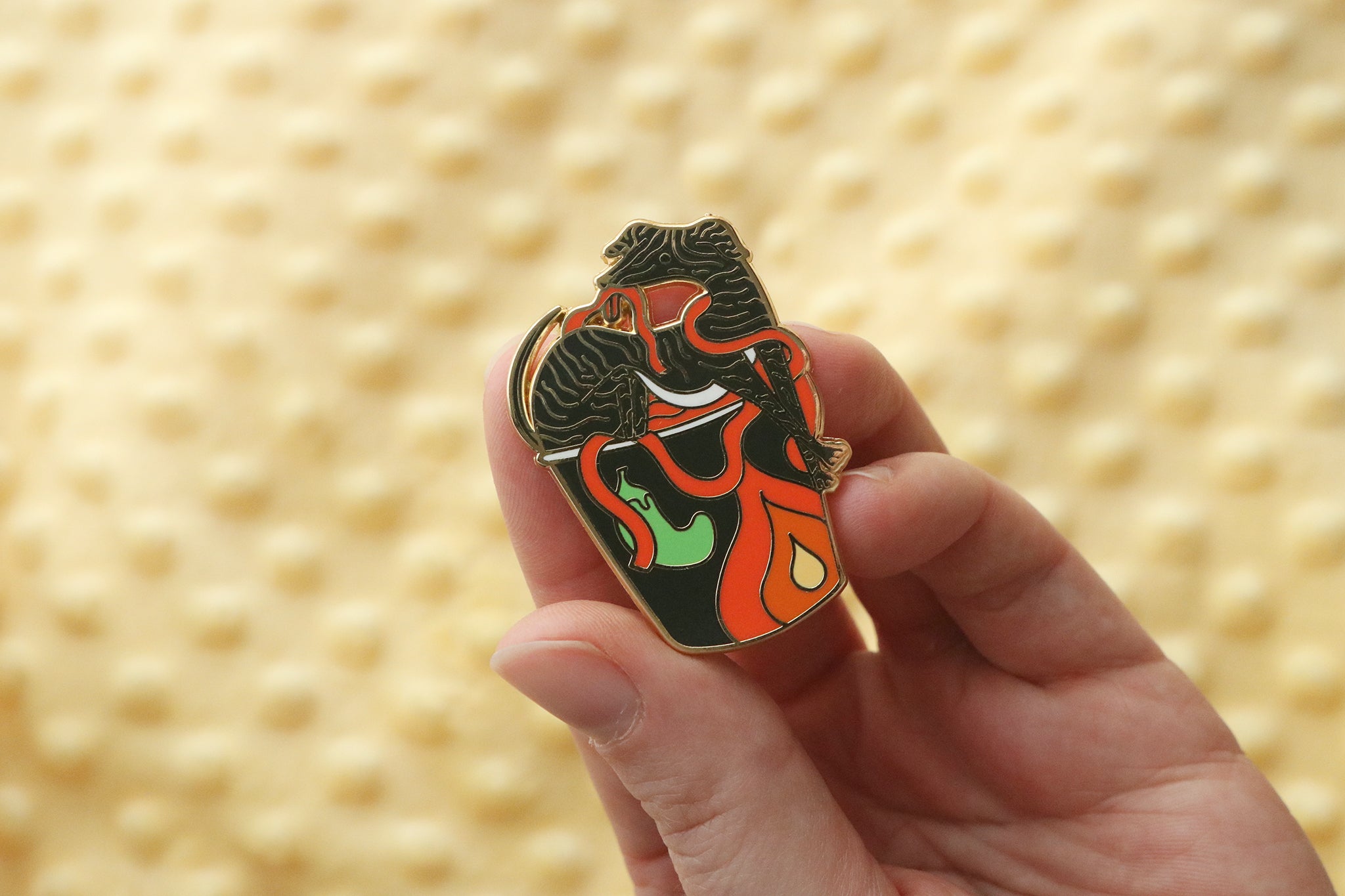 A gold plated enamel pin held in an open palm showing a stylized black hell hound emerging from a buldak noodle cup filled with wavy red spicy noodles.