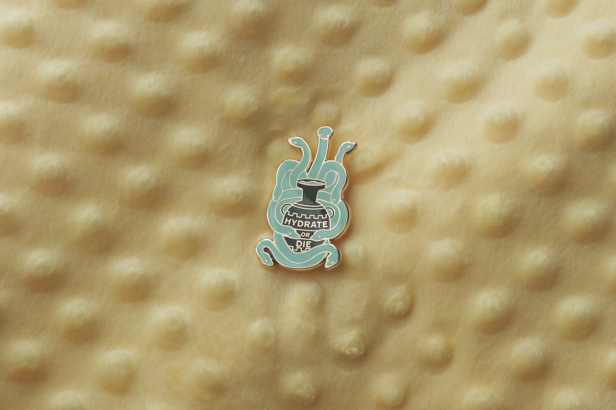 An enamel pin photographed against a yellow background, featuring a teal-colored hydra with multiple serpentine heads emerging from around a black ancient Greek amphora. The vessel displays text reading 'HYDRATE OR DIE' in gold plating.