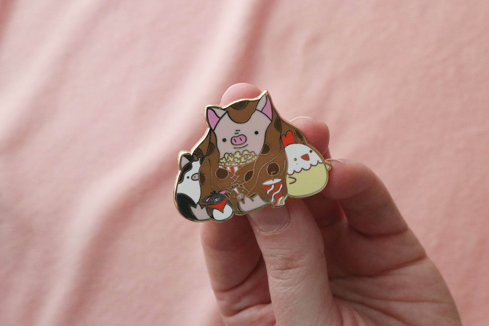 Hand holding an enamel pin of a cozy pig in a brown blanket eating popcorn, surrounded by small farm animal stuffed toys including a cow, donkey, horse, and chicken.