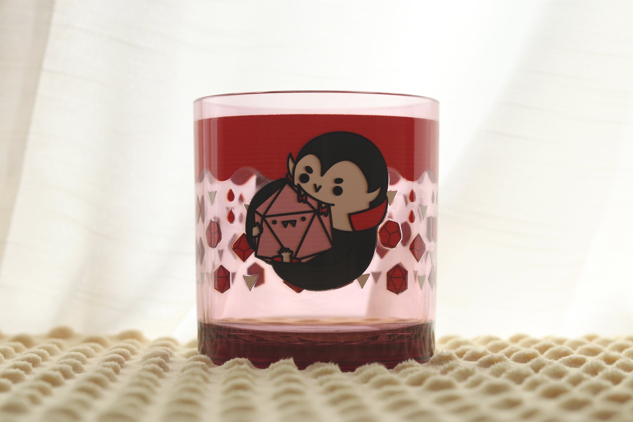 Blush colored drinking cup featuring a cute vampire character in black clothing sucking blood from a d20. Blood drips from the top of the glass and the vampire is surrounded by fangs and polyhedral dice.