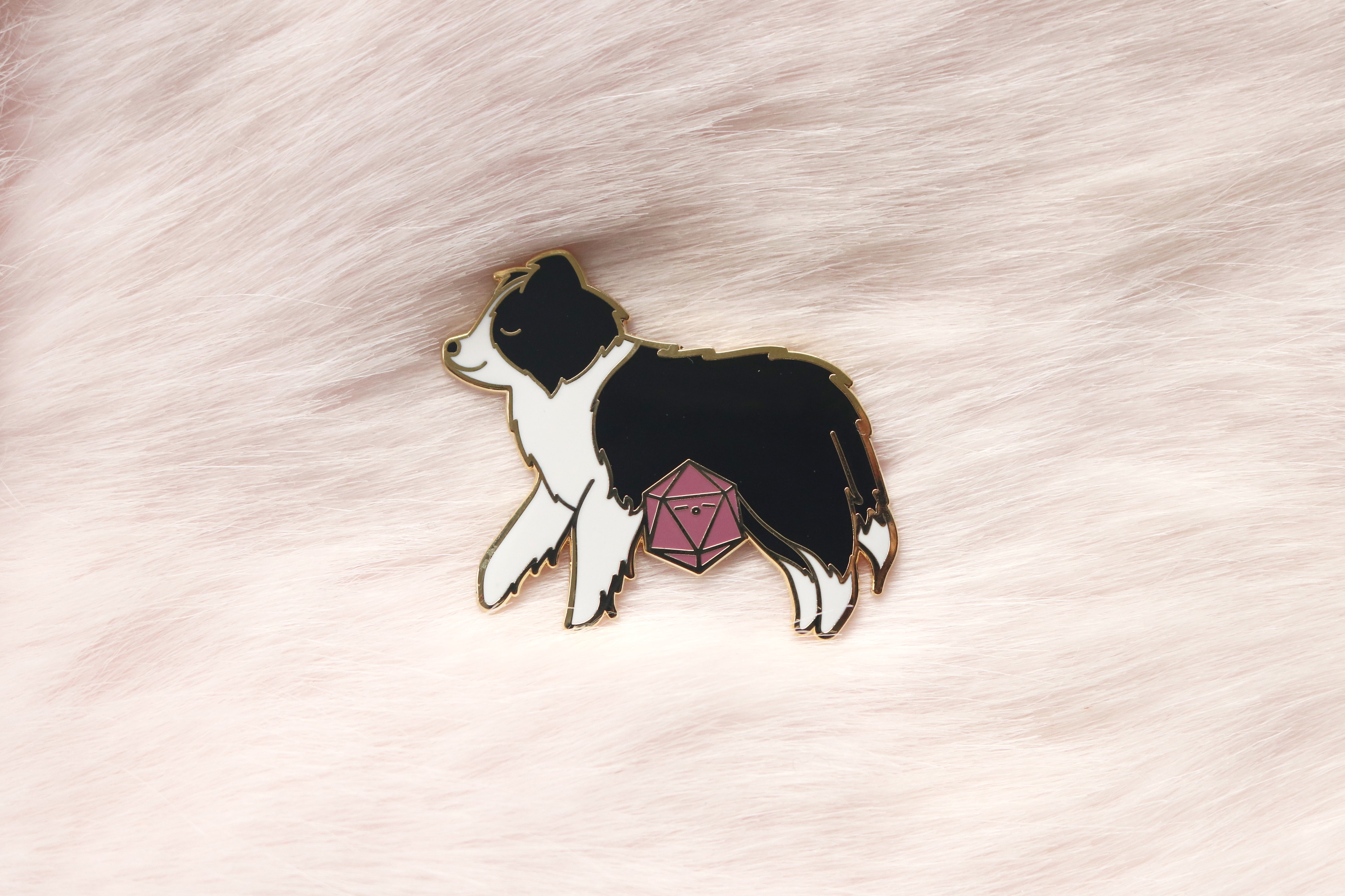 Enamel pin of a sleepy black and white Border Collie with a mauve colored d20. Photographed on light pink fur.