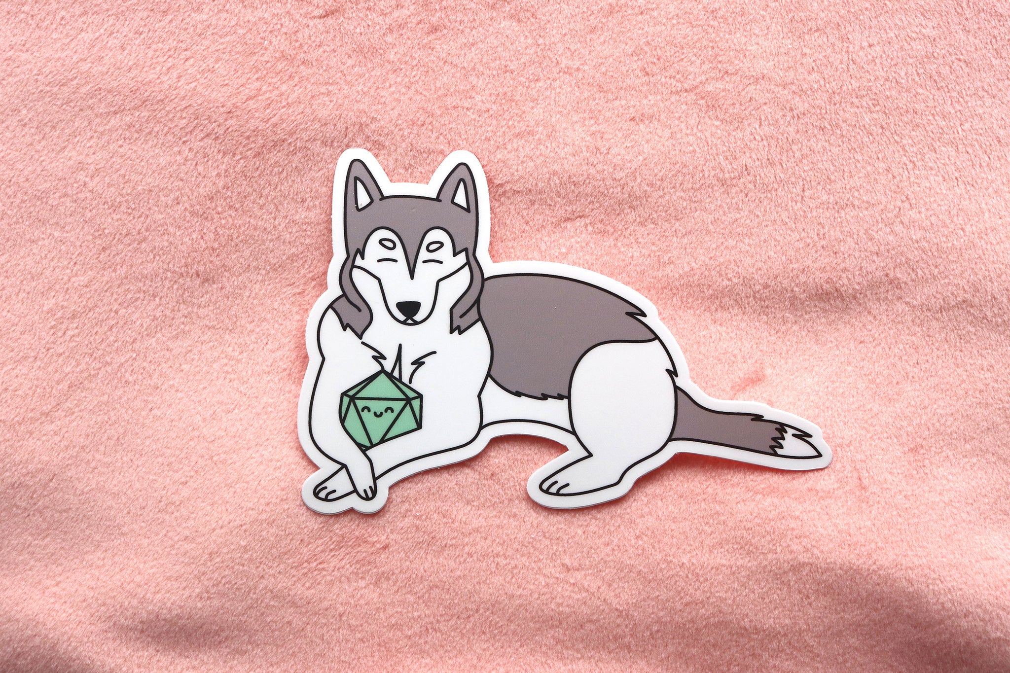 Gray and white husky sticker on pink fabric background. Dog is holding a mint colored d20 between its front paws. Both dog and dice have happy faces with closed eyes.